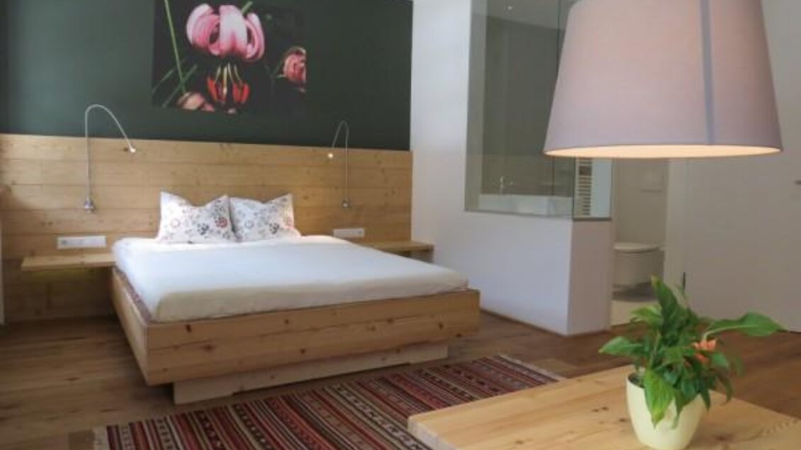 Ein modernes Schlafzimmer mit einem Holzbett und bunten Kissen. Die Wände sind in sanften Farben gehalten, und ein großer Lampenschirm sowie ein Pflanze auf dem Tisch sorgen für kuschelige Atmosphäre.