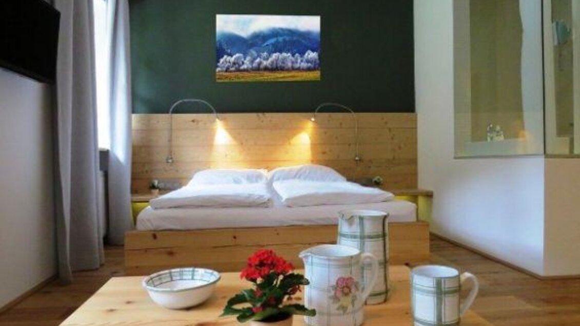 Ein gemütliches Schlafzimmer mit einem Holzbett und einem modernen Wandgemälde. Auf dem Tisch stehen eine Blume, eine Schüssel und zwei Kannen.