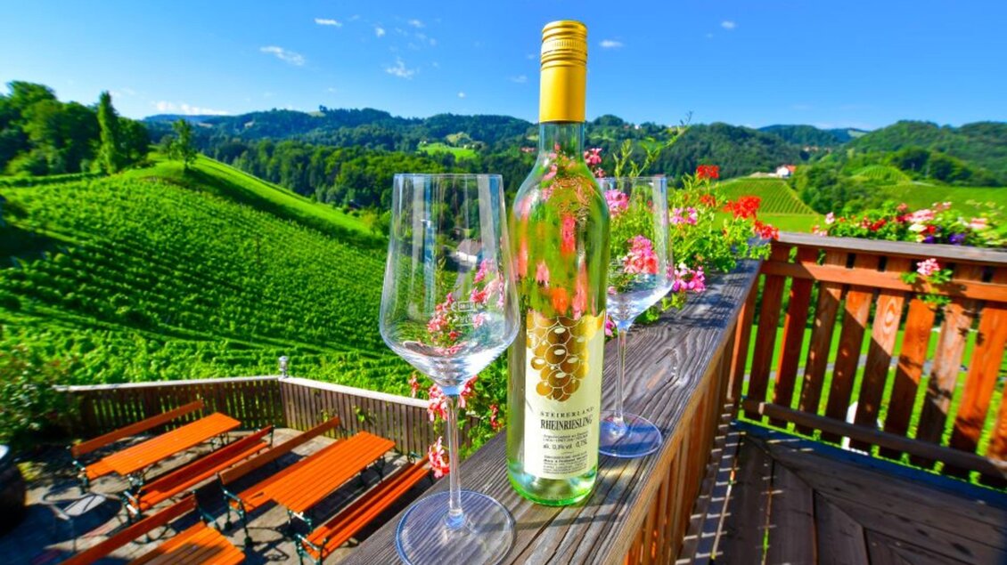 Ein schönes Weinberg mit einer Flasche Weißwein und Gläsern auf einem Holzgeländer. Im Hintergrund sind grünen Hügel und ein blauer Himmel zu sehen. | © Fam. Hernach | Der Hermannhof