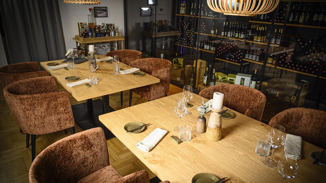 Ein stilvoll eingerichteter Restaurantbereich mit Holztischen und gemütlichen, samtigen Stühlen. Im Hintergrund ist eine gut sortierte Weinsammlung sichtbar. | © DI (FH) Philipp Wagner