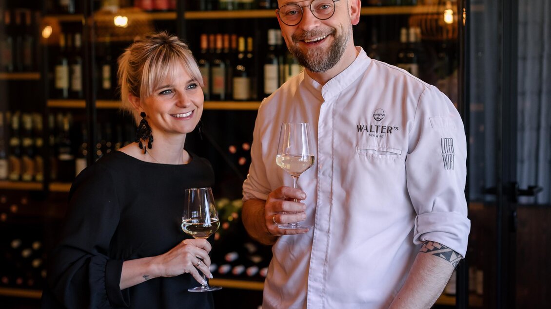 Ein Mann und eine Frau stehen lächelnd in einem stilvollen Restaurant. Beide halten ein Glas Getränke und die Atmosphäre ist einladend und gemütlich. | © Der Literwirt