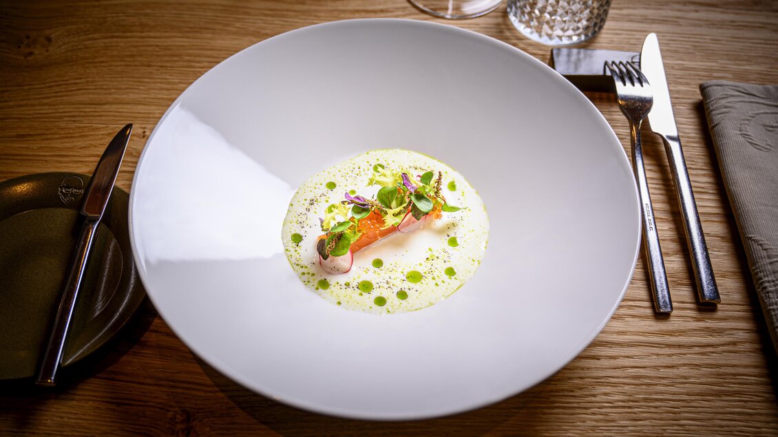 Ein elegantes Gericht in einem weißen Teller mit einer Portion Fisch und einer grünen Kräutersauce. Der Teller befindet sich auf einem holzernen Tisch. | © Der Literwirt