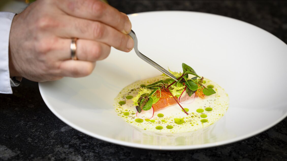 Ein Koch richtet ein elegantes Gericht auf einem weißen Teller an. Das Gericht besteht aus Lachs, einer cremigen Soße und frischen Kräutern. | © Der Literwirt