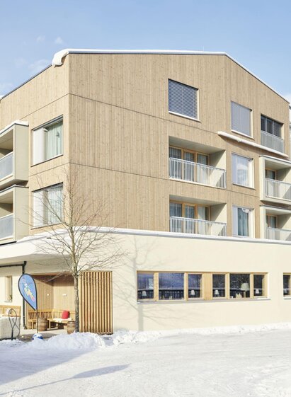 A modern building in winter with snow and mountains in the background. It is a hotel with large windows and an inviting entrance area. | © Stegerhof