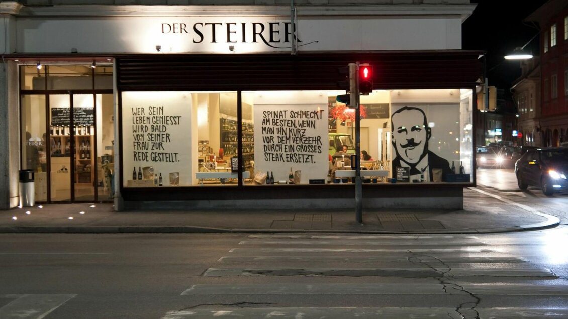 Ein Geschäft mit beleuchteter Fensterfront in der Nacht. An der Wand sind große Schriftzüge und ein Bild zu sehen. | © Weitzer