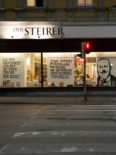 Ein Geschäft mit beleuchteter Fensterfront in der Nacht. An der Wand sind große Schriftzüge und ein Bild zu sehen. | © Weitzer