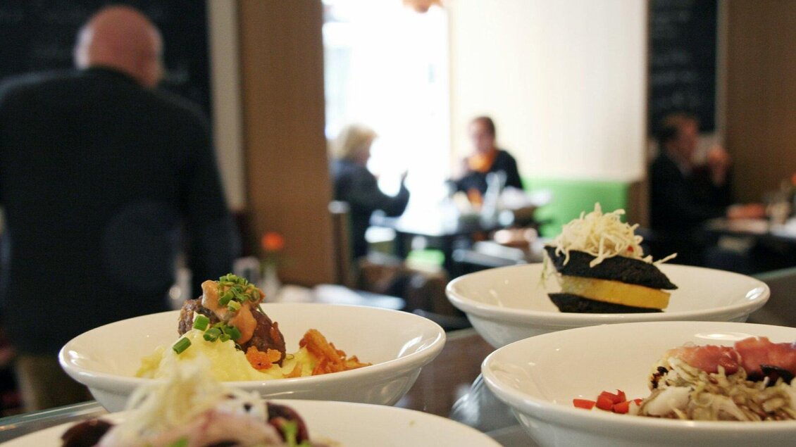 Ein Blick auf mehrere Schalen mit verschiedenen Gerichten in einem Restaurant. Im Hintergrund sind Gäste zu sehen, die sich unterhalten. | © Weitzer