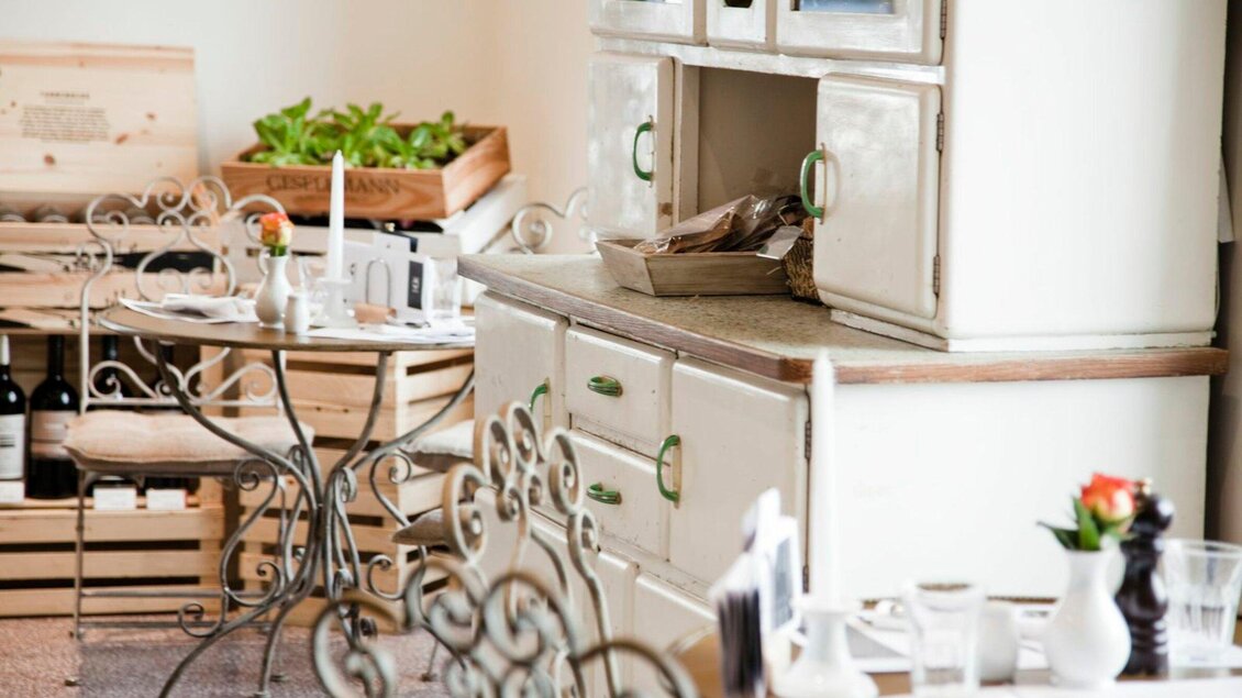 Eine gemütliche Innenansicht eines Restaurants mit einem weißen Schrank und dekorativen Tischen. Pflanzen und Speisen sind auf einem Regal im Hintergrund sichtbar. | © Weitzer