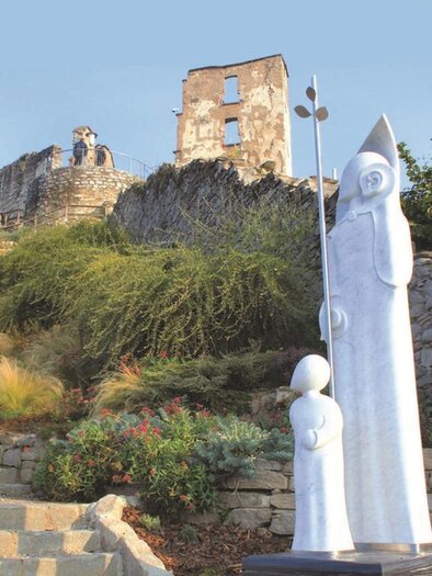 Eine moderne Skulptur zeigt eine Mutter mit ihrem Kind auf einer Treppe. Im Hintergrund sind die Ruinen einer alten burgartigen Struktur und grünes Gelände zu sehen. | © Robert Cescutti