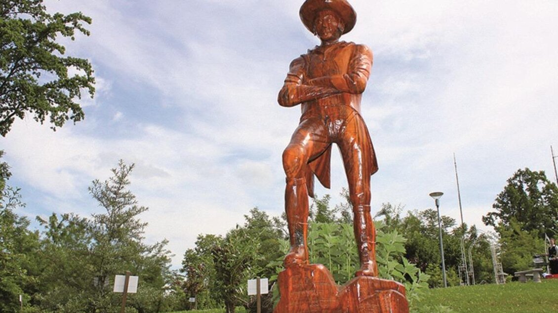 Eine große Holzstatue eines stehenden Mannes mit Hut, der selbstbewusst aussieht. Um ihn herum befinden sich grüne Bäume und Pflanzen. | © Robert Cescutti