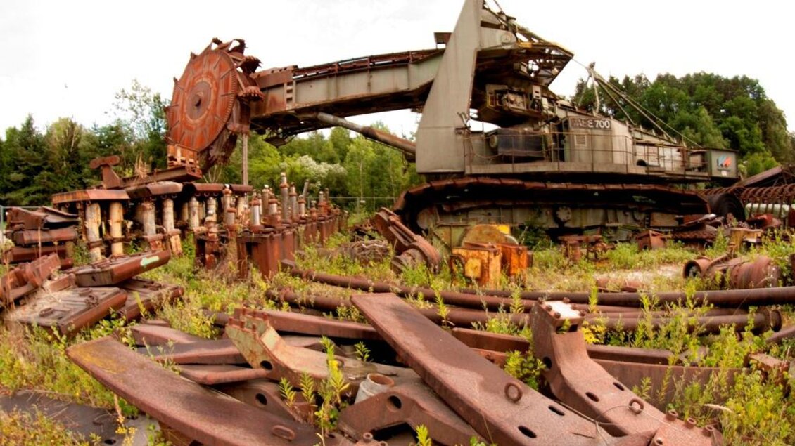 Eine alte, rostige Baggermaschine steht in einem verwilderten Industriegebiet. Umgeben von heruntergekommenen Metallteilen und Pflanzen zeigt die Szene einen verlassenen und vergessenen Ort. | © Heimo Kos