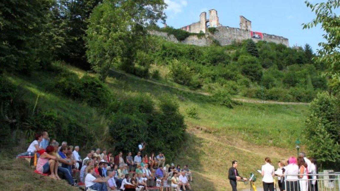 Eine Gruppe von Menschen sitzt auf einer Wiese vor einer alten Burg. Der Himmel ist klar und das Gelände ist grün und bewaldet. | © Keine Kenntnis