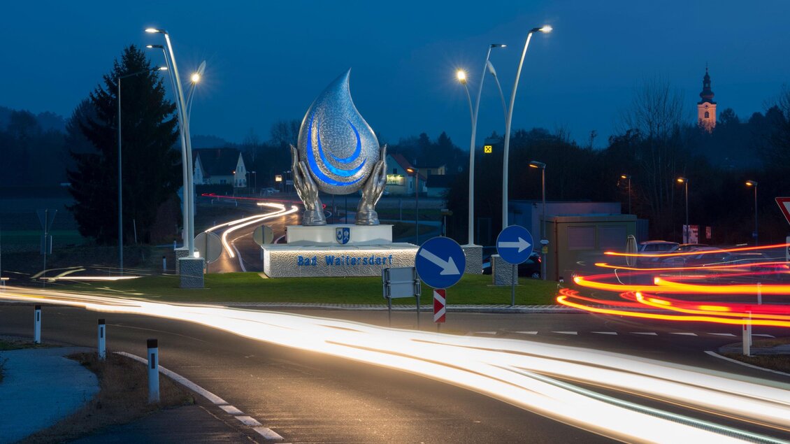 Ein beleuchtetes Denkmal an einem Kreisverkehr bei Nacht. Der Himmel ist dunkel und es sind Lichtspuren von Autos sichtbar. | © Bernhard Bergmann