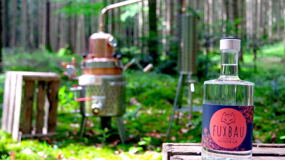 Eine Flasche Fuxbau Spirituose steht auf einer Holzpalette im Wald. Im Hintergrund befindet sich eine Kupferdestillieranlage zwischen den Bäumen. | © Martin Strasser
