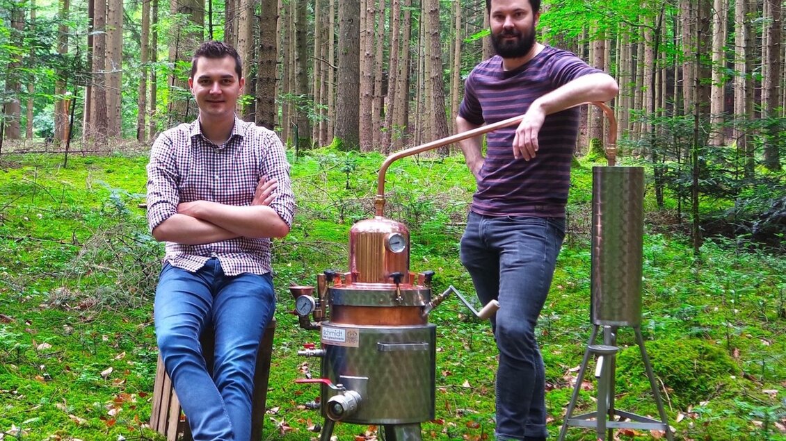 Zwei Männer stehen im Wald neben einer kleinen Destillieranlage. Der Wald wirkt grün und idyllisch, mit hohen Bäumen im Hintergrund. | © Martin Strasser