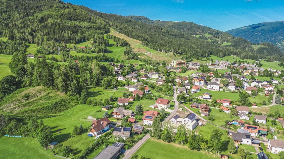 Eine malerische Aussicht auf ein kleines Dorf umgeben von grünen Hügeln und Bergen. Der Himmel ist klar und blau, was die idyllische Landschaft unterstreicht. | © Tourismusverband Murau