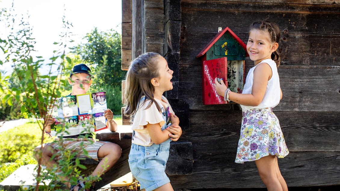 Zwei fröhliche Mädchen stehen vor einer Holzhütte und lachen. Ein Junge sitzt im Hintergrund und hält ein Heft in der Hand. | © TVB Murau