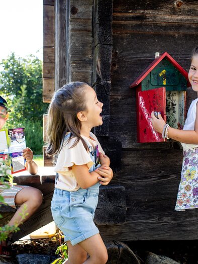 Two cheerful girls stand in front of a wooden hut and laugh. A boy is sitting in the background holding a notebook in his hand. | © TVB Murau