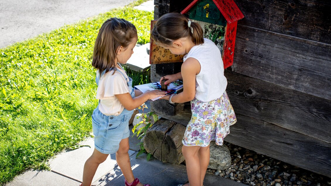 Zwei Kinder stehen draußen und schauen sich etwas an. Im Hintergrund ist ein Holzhaus mit einer roten Briefkasten. | © TVB Murau