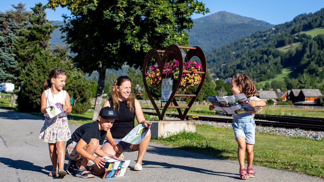 Eine Familie mit zwei Kindern steht auf einer Straße und schaut auf Karten. Im Hintergrund sind grüne Berge und ein Herz aus Blumen zu sehen. | © TVB Murau