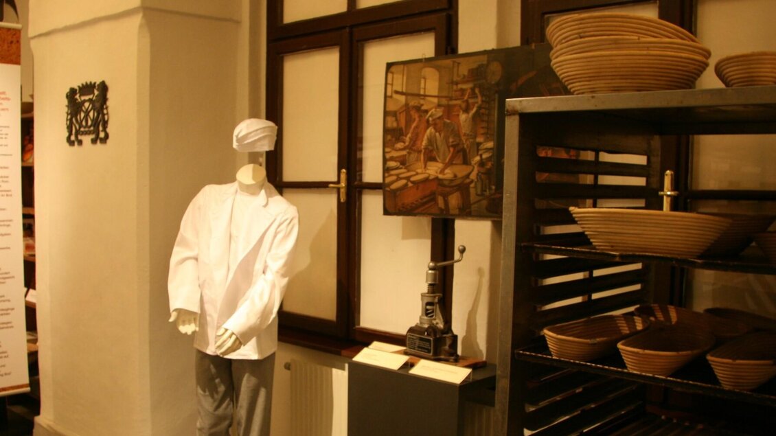 Exponate zur Brotbackkunst mit Bäckerkleidung, Gärkorb und Infotafel im Museum | © Diözesanmuseum Graz