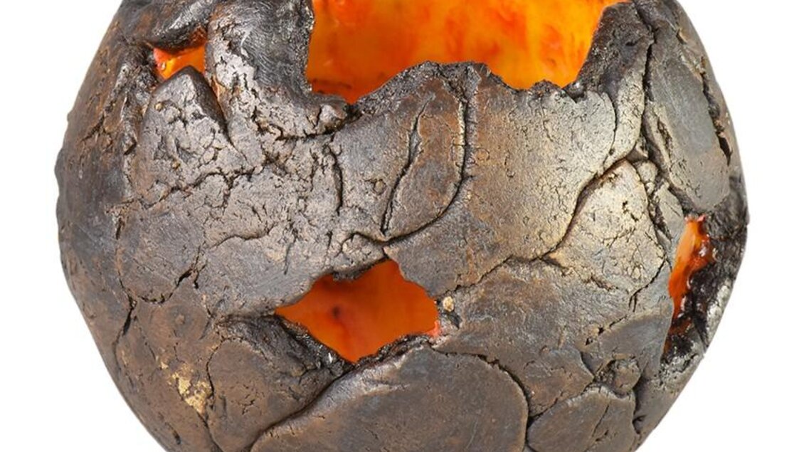 Eine strukturierte Schale mit einer dunklen, rauen Oberfläche und einem leuchtend orangefarbenen Inneren. Sie hat unregelmäßige Löcher und sieht aus wie aus Lava oder Ton gefertigt.