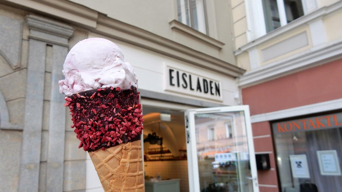 Ein leckeres Eis in einer Waffel vor einem Eisladen. Die Waffel hat eine rosa Eiskugel und ist mit bunten Streuseln dekoriert. | © Stadtmarketing Judenburg