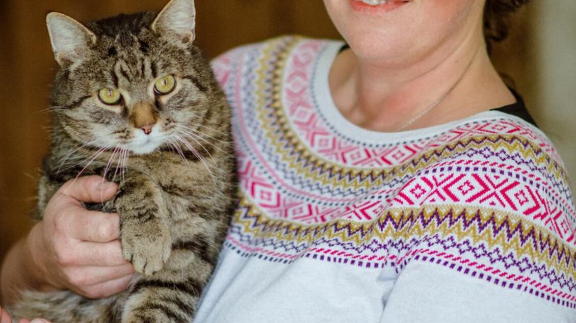 Eine Frau hält eine getigerte Katze in ihren Armen und lächelt freundlich. Sie trägt ein auffälliges Sweatshirt mit buntem Muster. | © Christoph Huber