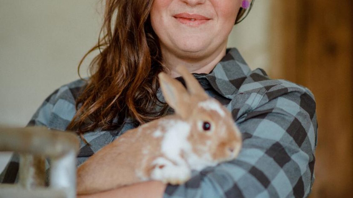 Eine Frau hält ein Kaninchen in ihren Armen und lächelt glücklich. Sie trägt ein kariertes Hemd und steht in einem Stall. | © Christoph Huber