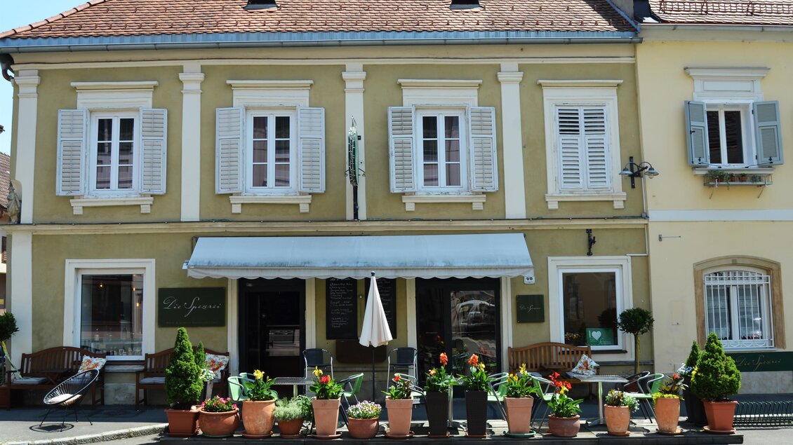 Ein charmantes Gebäude mit einem schattigen Vordach und großen Fenstern. Vor dem Gebäude blühen farbige Pflanzen in Töpfen. | © Die Spezerei