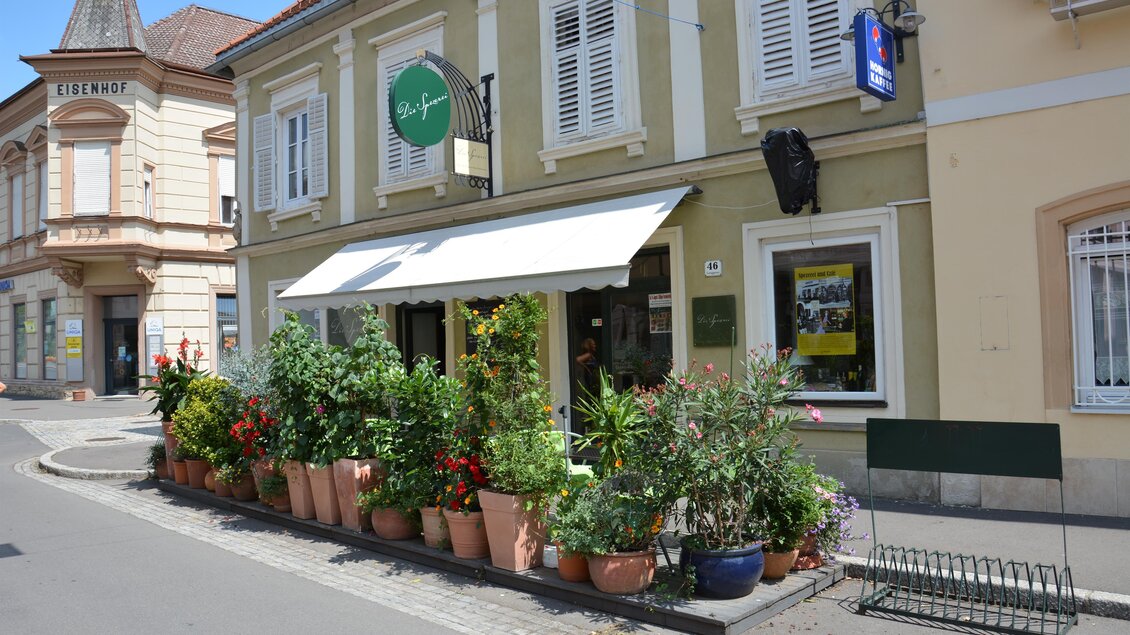 Ein einladendes Café an einer Straßenecke mit blühenden Pflanzen in Töpfen. Die Fassade des Gebäudes ist hell und freundlich. | © Hermann