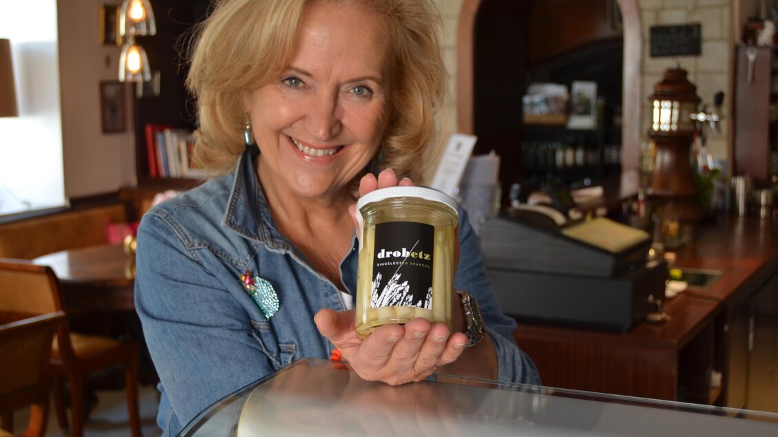 Eine Frau hält ein Glas eingelegter Produkte in der Hand und lächelt erfreut. Im Hintergrund ist eine gemütliche Café-Umgebung zu sehen. | © Die Spezerei