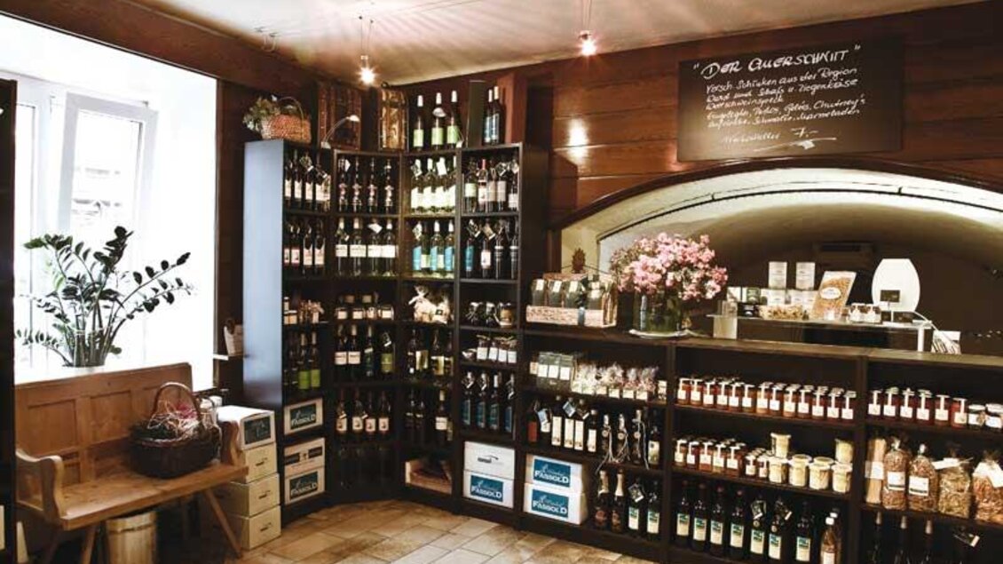 Ein gemütlicher Laden mit Regalen voller Weinflaschen und verschiedenen Produkten. Auf der Theke stehen frische Blumen und es gibt eine einladende Atmosphäre.