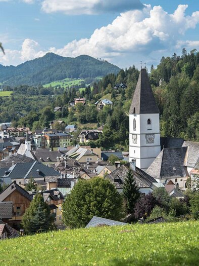 A picturesque town with traditional buildings and a striking church. In the background, green hills and mountains can be seen. | © Herbert Sams