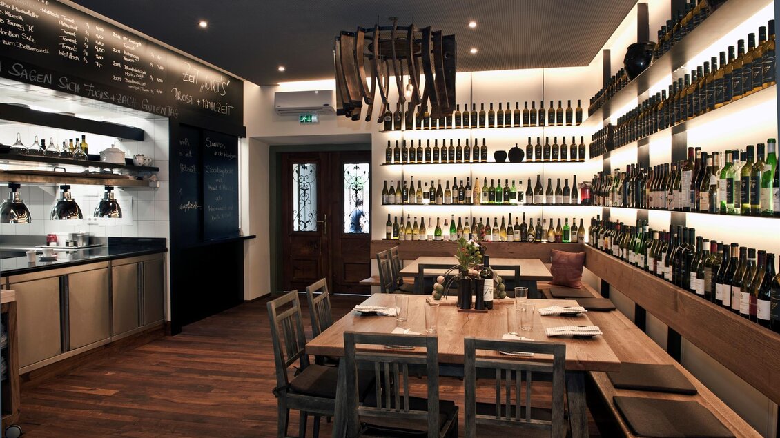 Ein stilvolles Restaurant mit Holztischen und einer Wand voller Weinflaschen. Die Beleuchtung schafft eine gemütliche Atmosphäre. | © Breitenthaler