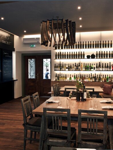 A stylish restaurant with wooden tables and a wall full of wine bottles. The lighting creates a cozy atmosphere. | © Breitenthaler