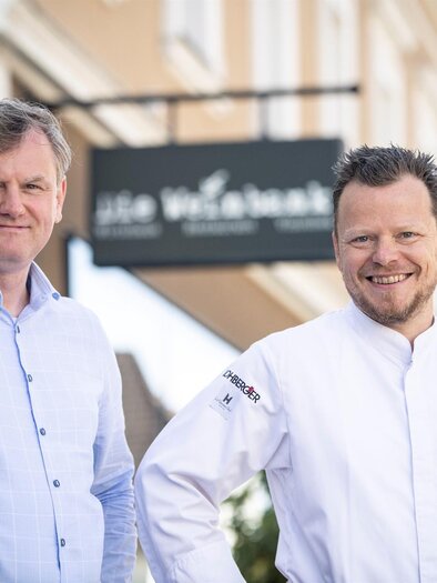 Two men are smiling in front of a restaurant. In the background, a sign with the name of the establishment is visible. | © Die Weinbank | Christian Jungwirth