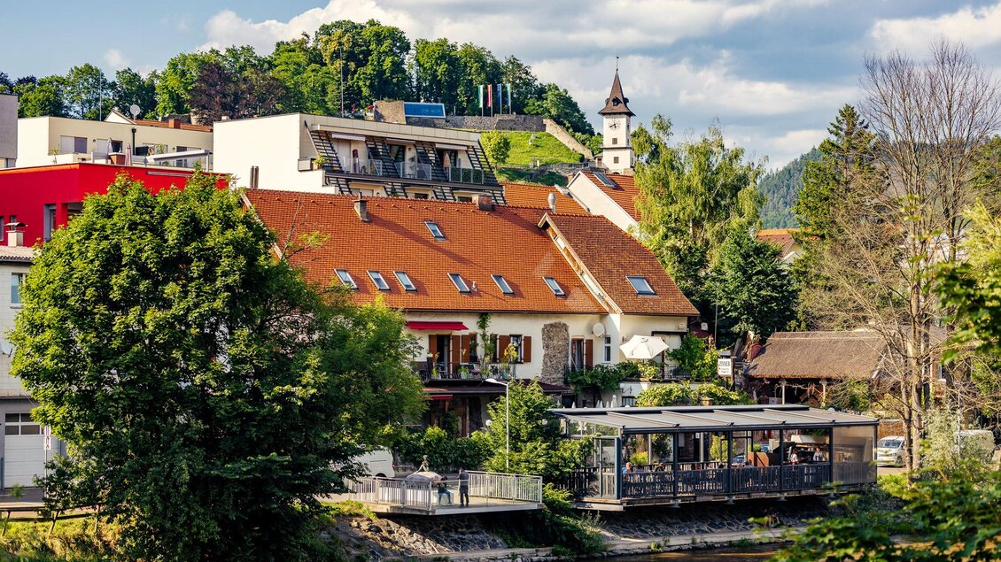Eine malerische Stadtansicht mit bunten Häusern und grünen Bäumen. Im Hintergrund steht ein Kirchturm und fließt ein ruhiger Fluss. | © TV Hochsteiermark