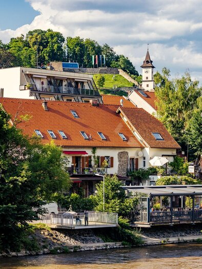 A picturesque town view with colorful houses and green trees. In the background, there is a church tower and a calm river flows. | © TV Hochsteiermark