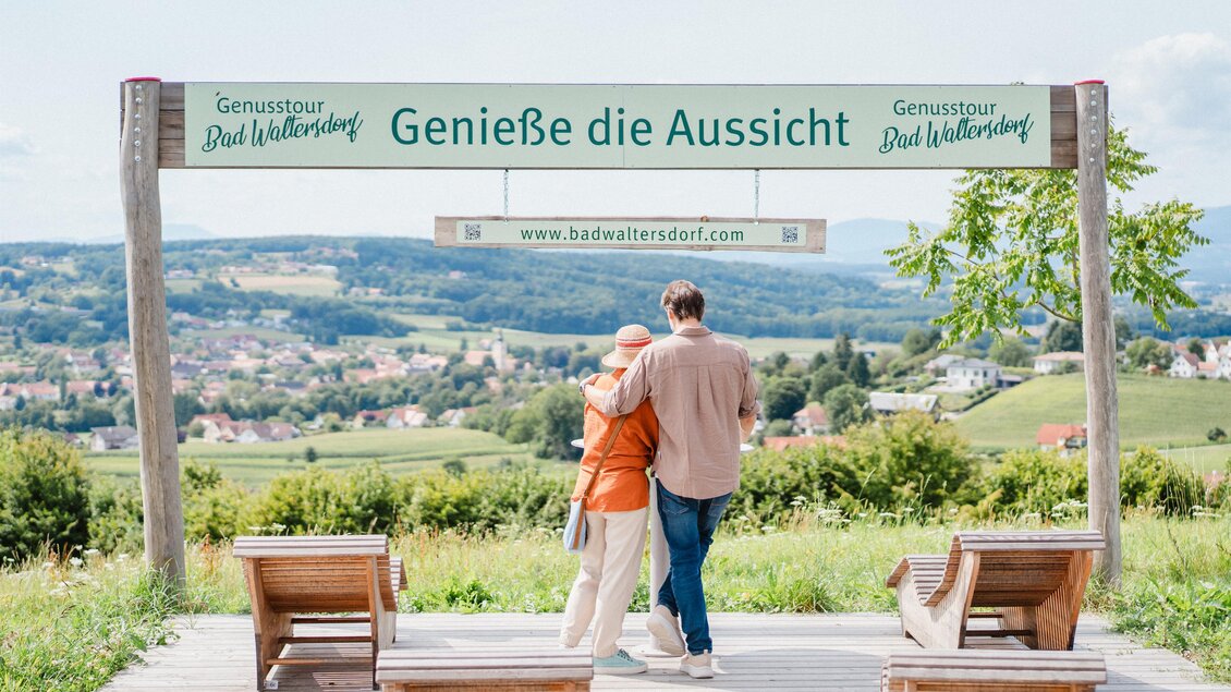 Zwei Personen stehen auf einer Aussichtsplattform mit Blick auf eine malerische Landschaft. Ein Schild lädt dazu ein, die Aussicht zu genießen. | © motionads.at_KK Bad Waltersdorf