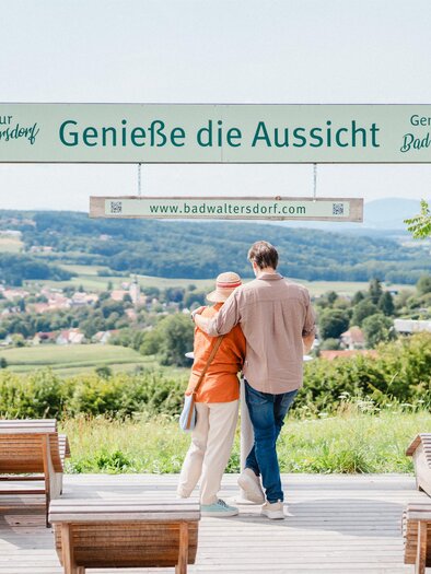 Two people are standing on an observation deck overlooking a picturesque landscape. A sign invites them to enjoy the view. | © motionads.at_KK Bad Waltersdorf