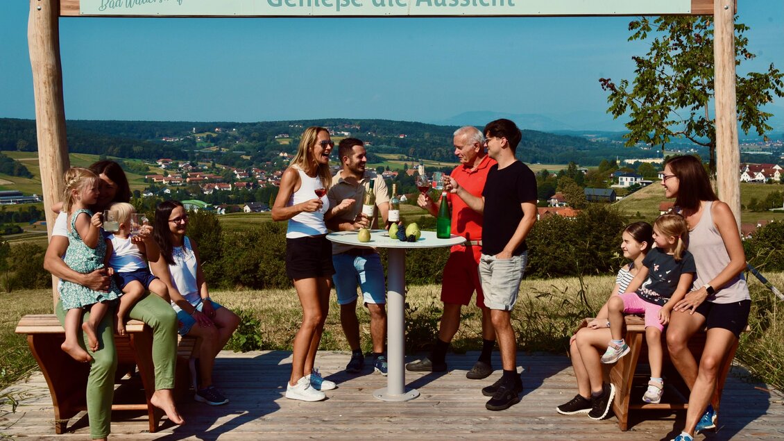 Eine fröhliche Gruppe von Menschen genießt die Aussicht an einem Tisch im Freien. Sie lachen und haben Getränke, umgeben von grünen Wiesen und einem weiten Blick auf die Landschaft. | © GDE Bad Waltersdorf