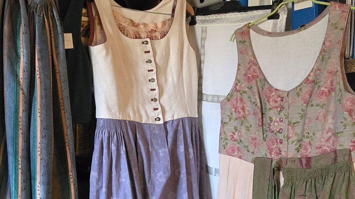 Traditionelle Kleider hängen an einer Wand. Die Outfits sind in verschiedenen Farben und Mustern, darunter florale und einfarbige Designs. | ©  Stephanie Bor