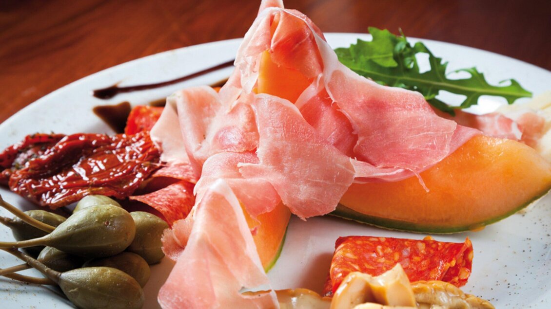 Ein Teller mit melonenförmigem Obst und hauchdünnem Schinken. Daneben liegen Kapern und getrocknete Tomaten. | © Don Camillo