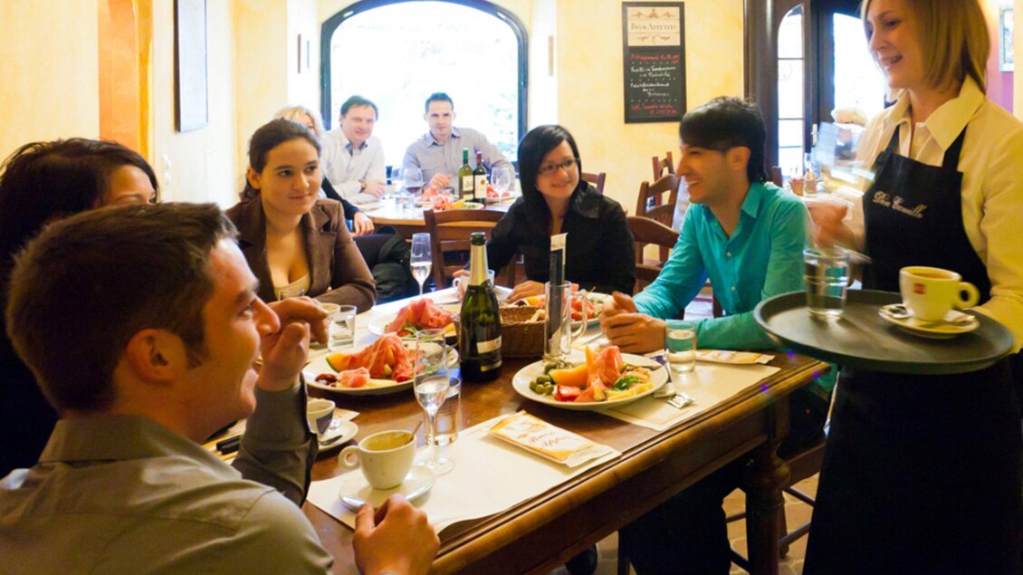 Eine Gruppe von Menschen sitzt an einem Tisch in einem Restaurant und genießt ihr Essen. Eine Kellnerin serviert Getränke und Speisen. | © Don Camillo
