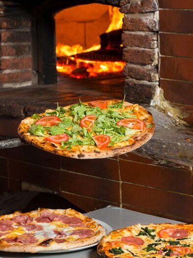 Ein Holzofen mit einem Pizzabäcker, der eine frische Pizza mit Tomaten und Rucola herausnimmt. Im Vordergrund sind zwei weitere Pizzen mit verschiedenen Belägen zu sehen. | © TV Region Graz Rene Vidalli.