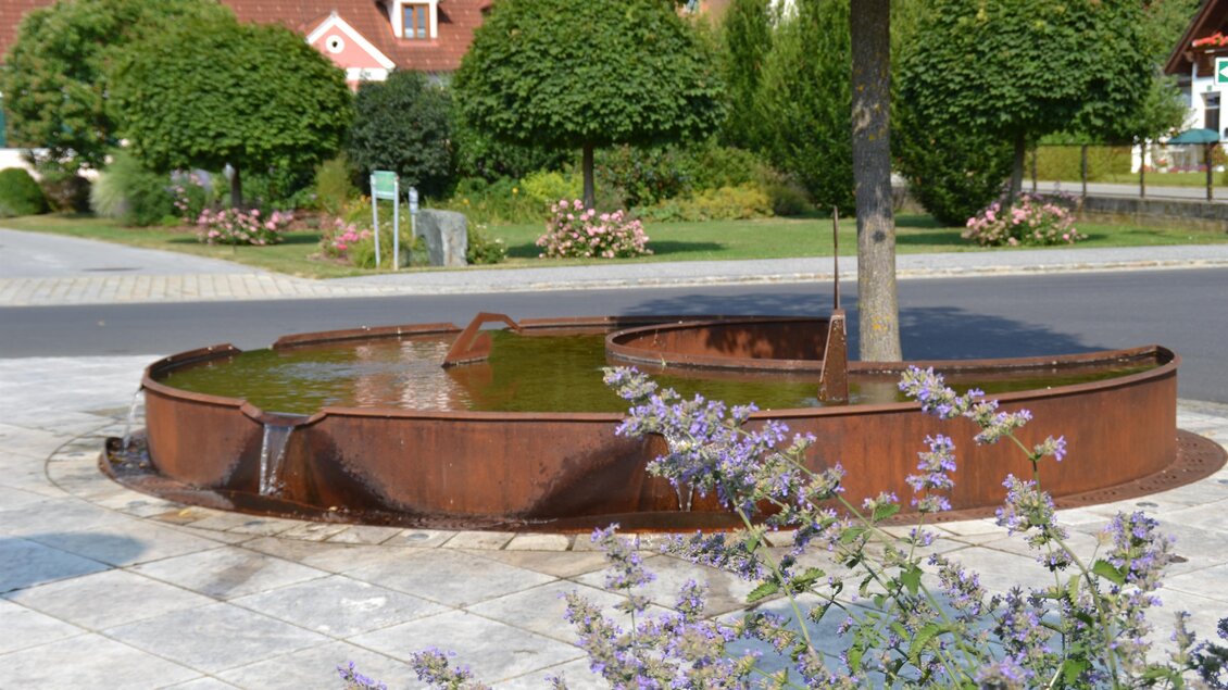 Eine moderne Brunnenanlage umgeben von Bäumen und Blumen. Das Wasser fließt sanft in eine runde, metallene Schale. | © Kurkommission Bad Blumau