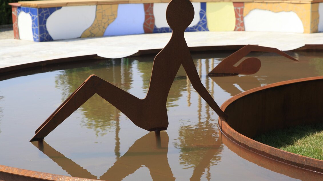 Eine moderne Skulptur in einem Wasserbecken, die eine sitzende Figur darstellt. Im Hintergrund sind bunte Fliesen sichtbar. | © Kurkommission Bad Blumau