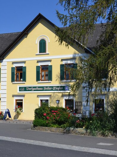 A yellow building with green window frames and old architecture. In front of the house, colorful flowers and trees can be seen. | © Riedenbauer