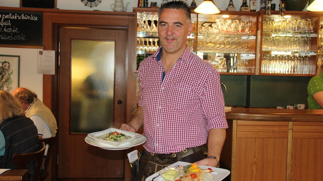 Ein Kellner trägt zwei Teller mit verschiedenen Gerichten in einem Restaurant. Im Hintergrund sind Gäste und eine gut ausgestattete Bar zu sehen. | © Riedenbauer